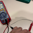 A red and black digital multimeter on a light beige surface, with a hand holding red test leads connected to its probes.