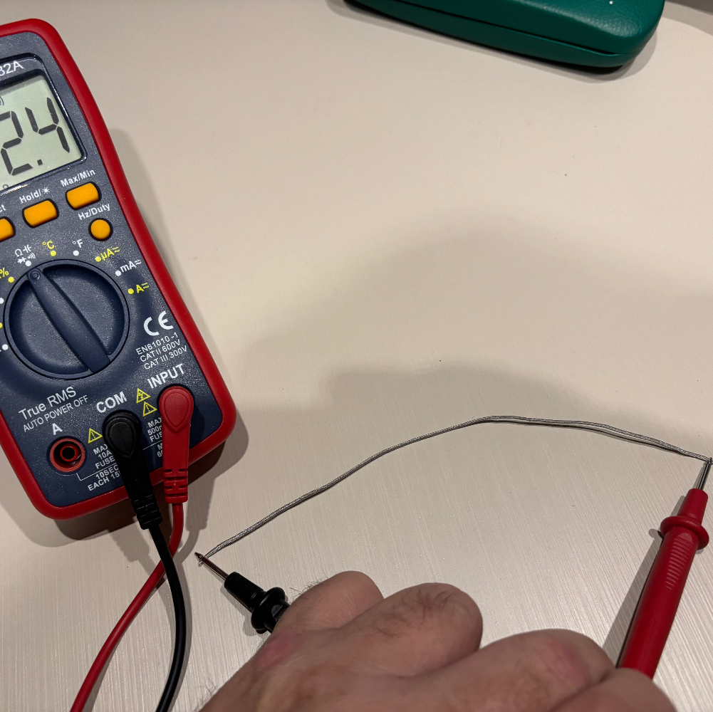 A red and black digital multimeter on a light beige surface, with a hand holding red test leads connected to its probes.