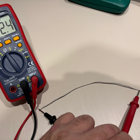A red and black digital multimeter on a light beige surface, with a hand holding red test leads connected to its probes.