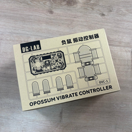 A beige product box for the DC-LAB Opossum Vibrate Controller (OVC-1) lies on a light-colored wood floor. The box features black text and technical line drawings of the controller.